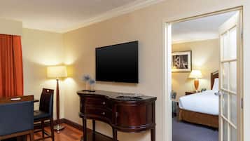 Habitación de hotel con televisor de pantalla plana montado en la pared, escritorio con lámpara, silla y cama con cubrecama blanco.