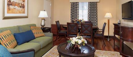 A hotel room with a green sofa, a dining table with chairs, a coffee table with flowers, a TV, and a painting on the wall.