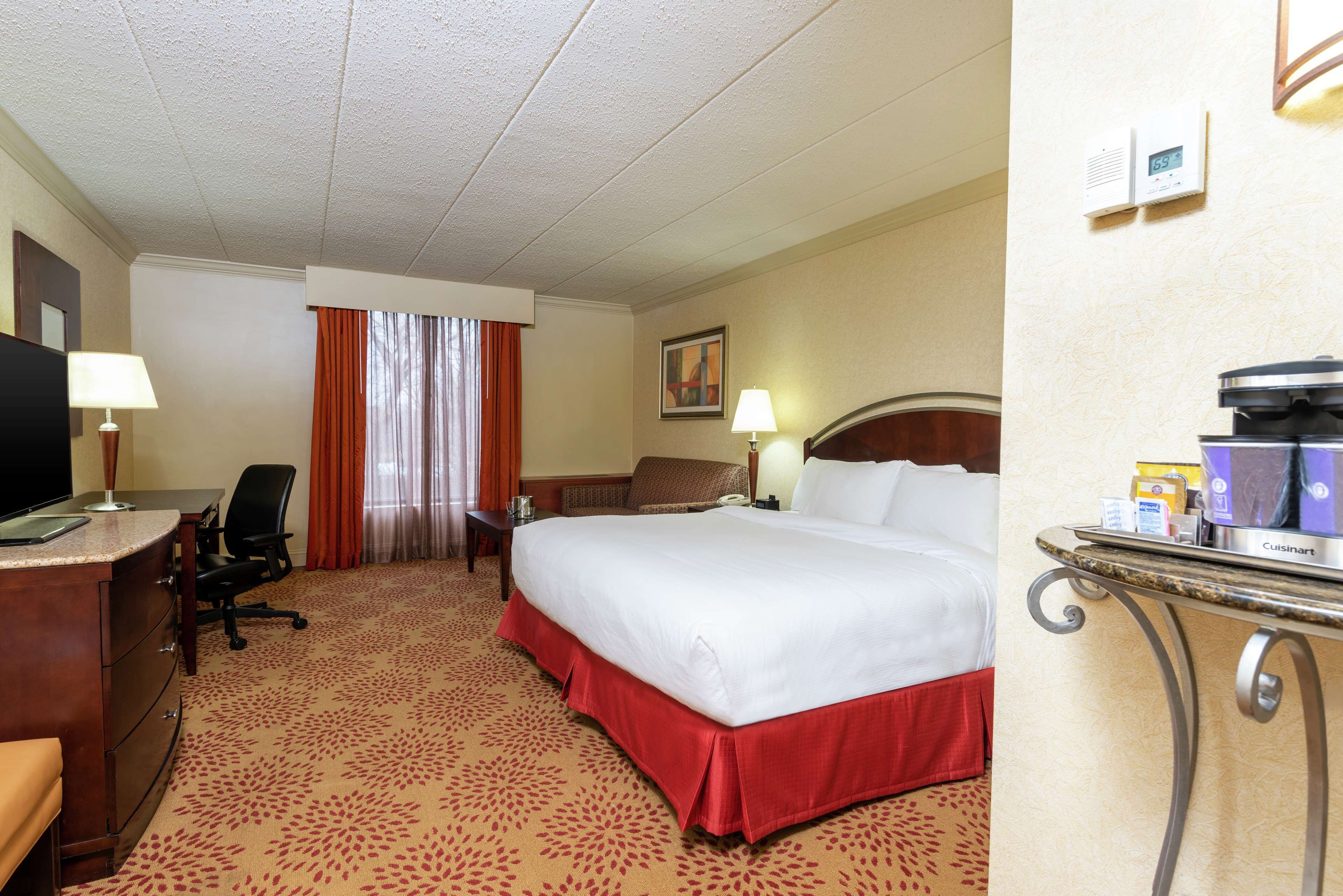 A hotel room with a large bed, a desk with a computer, a chair, a TV, and a window with curtains.