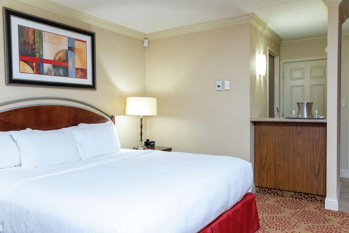 A hotel room with a large bed, a nightstand with a lamp, a framed abstract painting, and a wooden cabinet.