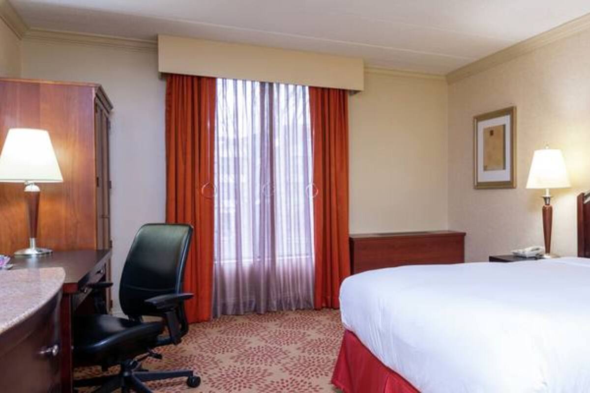 A hotel room with a bed, a desk, a chair, a lamp, and a window with curtains.