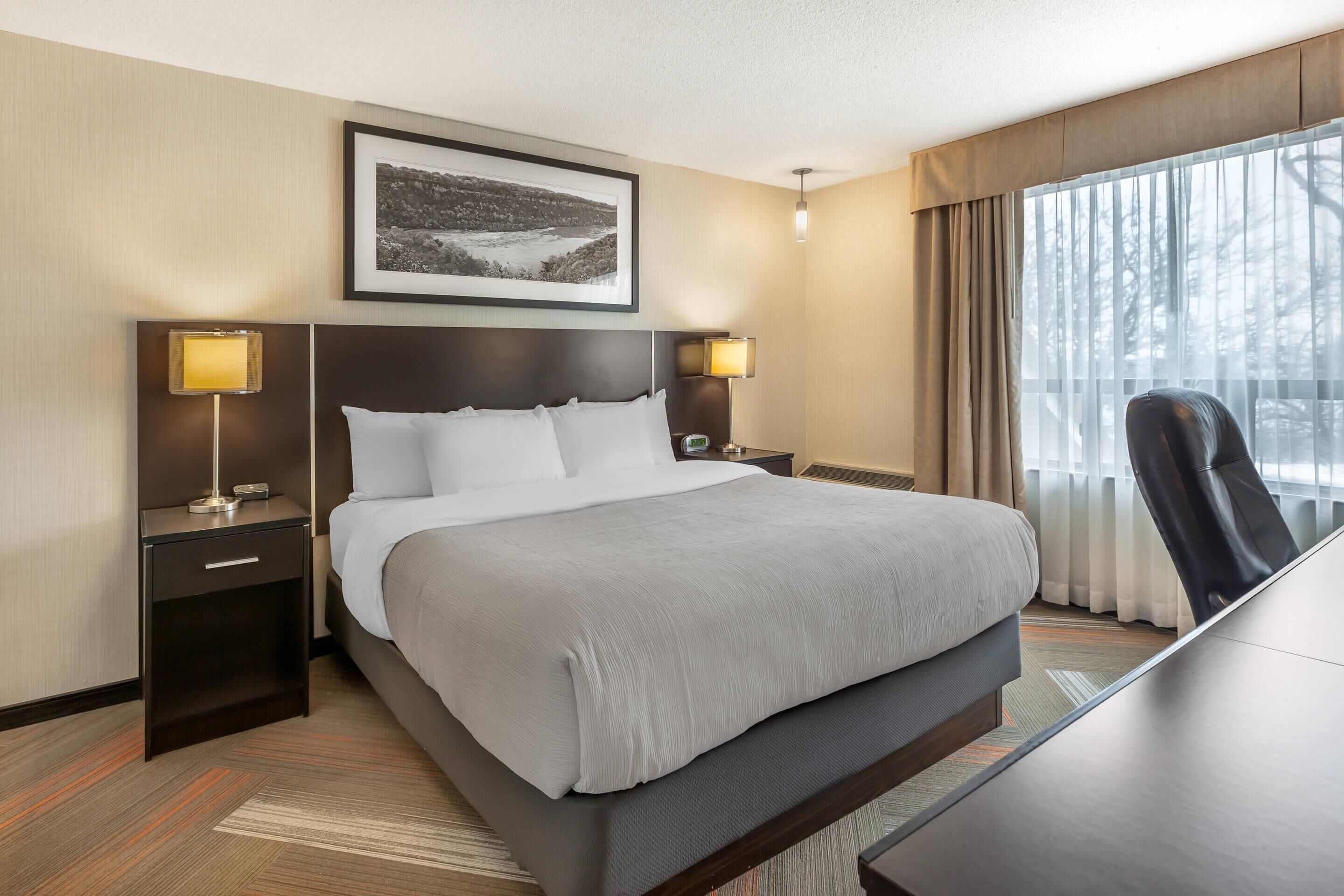 Quarto de hotel com uma cama grande, luminárias de cabeceira, uma escrivaninha e uma janela com cortinas.