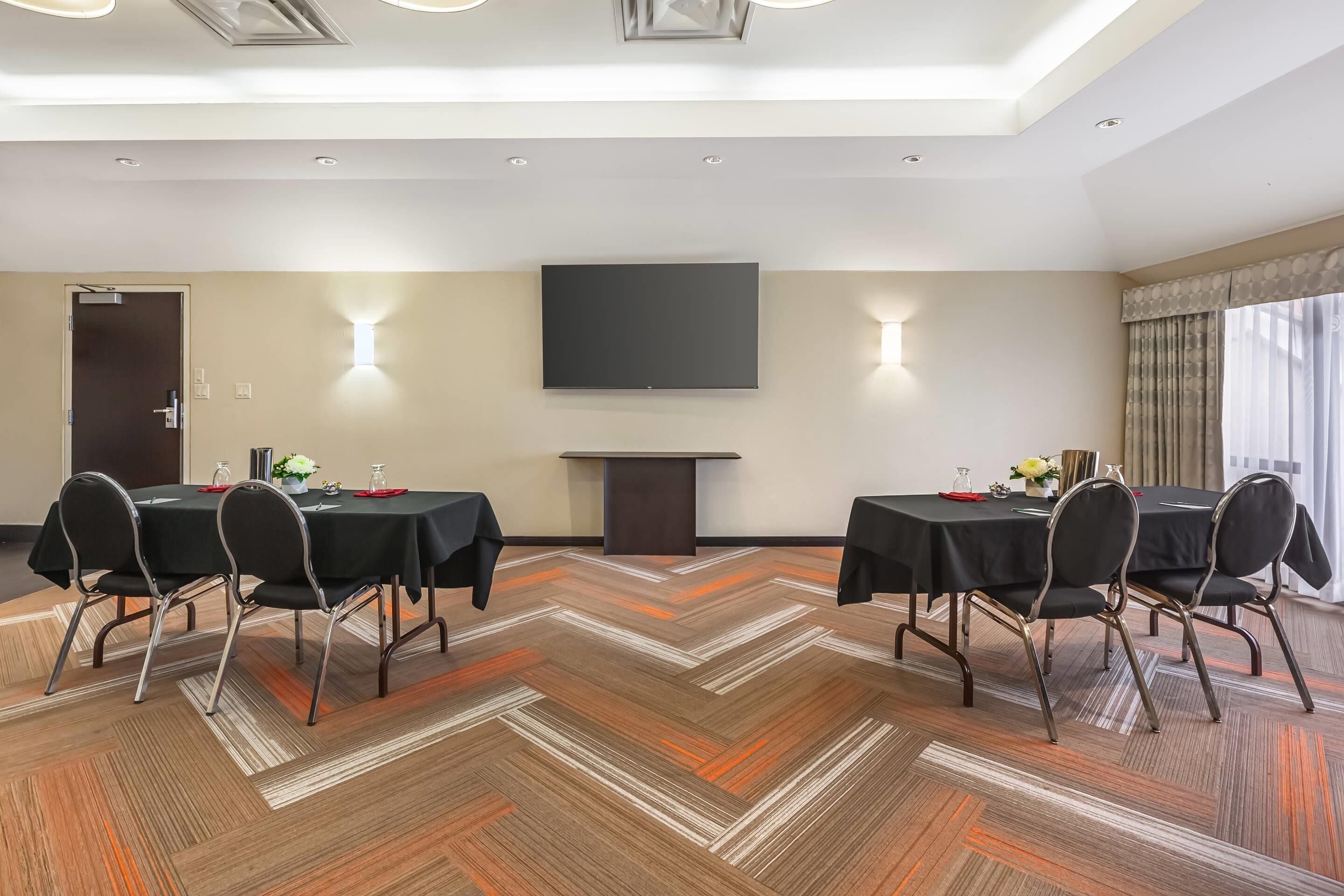 A conference room with a large flat-screen TV, two tables with chairs, a patterned carpet, and a door.