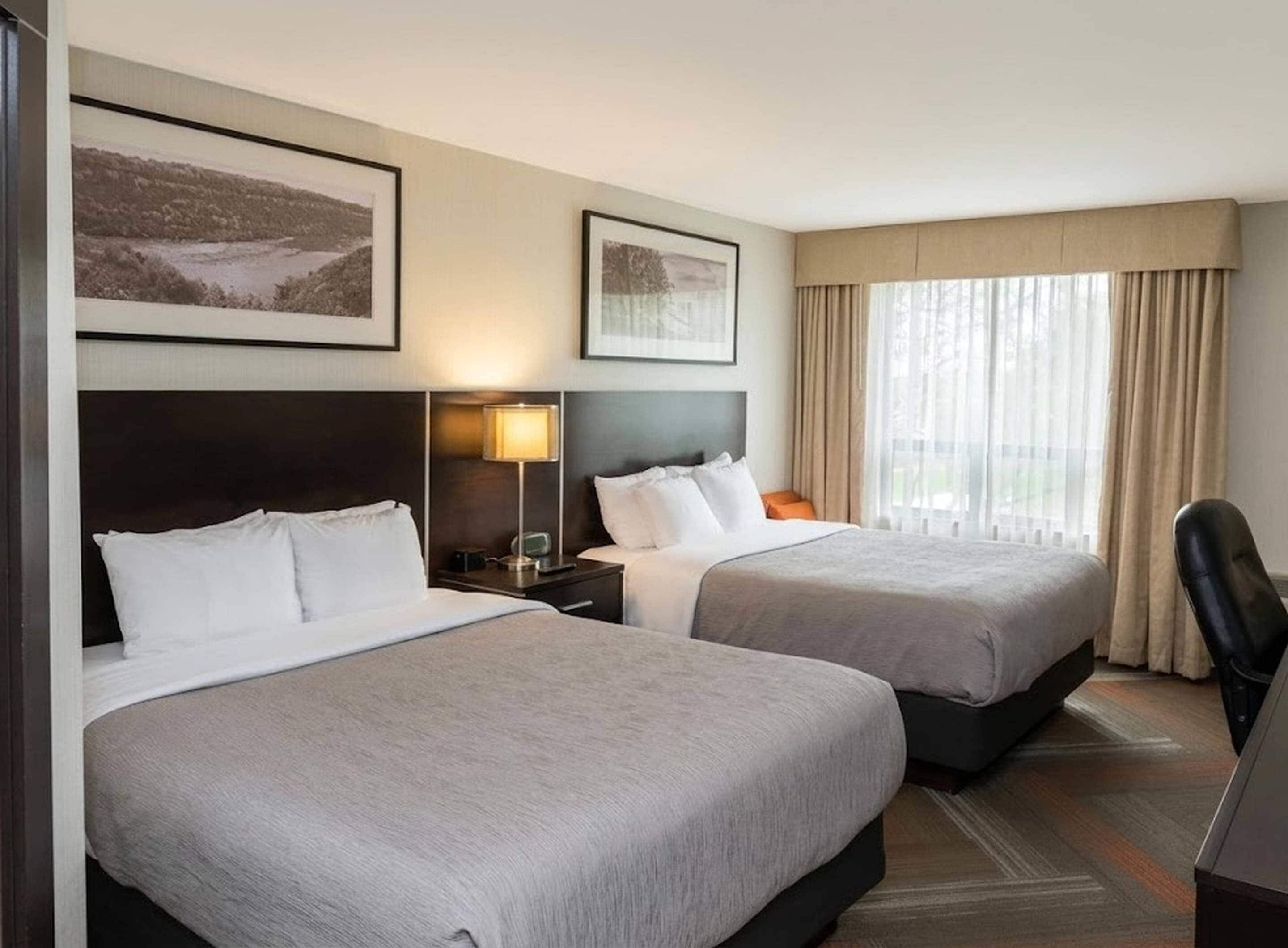 A hotel room with two beds, a nightstand with a lamp, a chair, and two framed pictures on the wall.