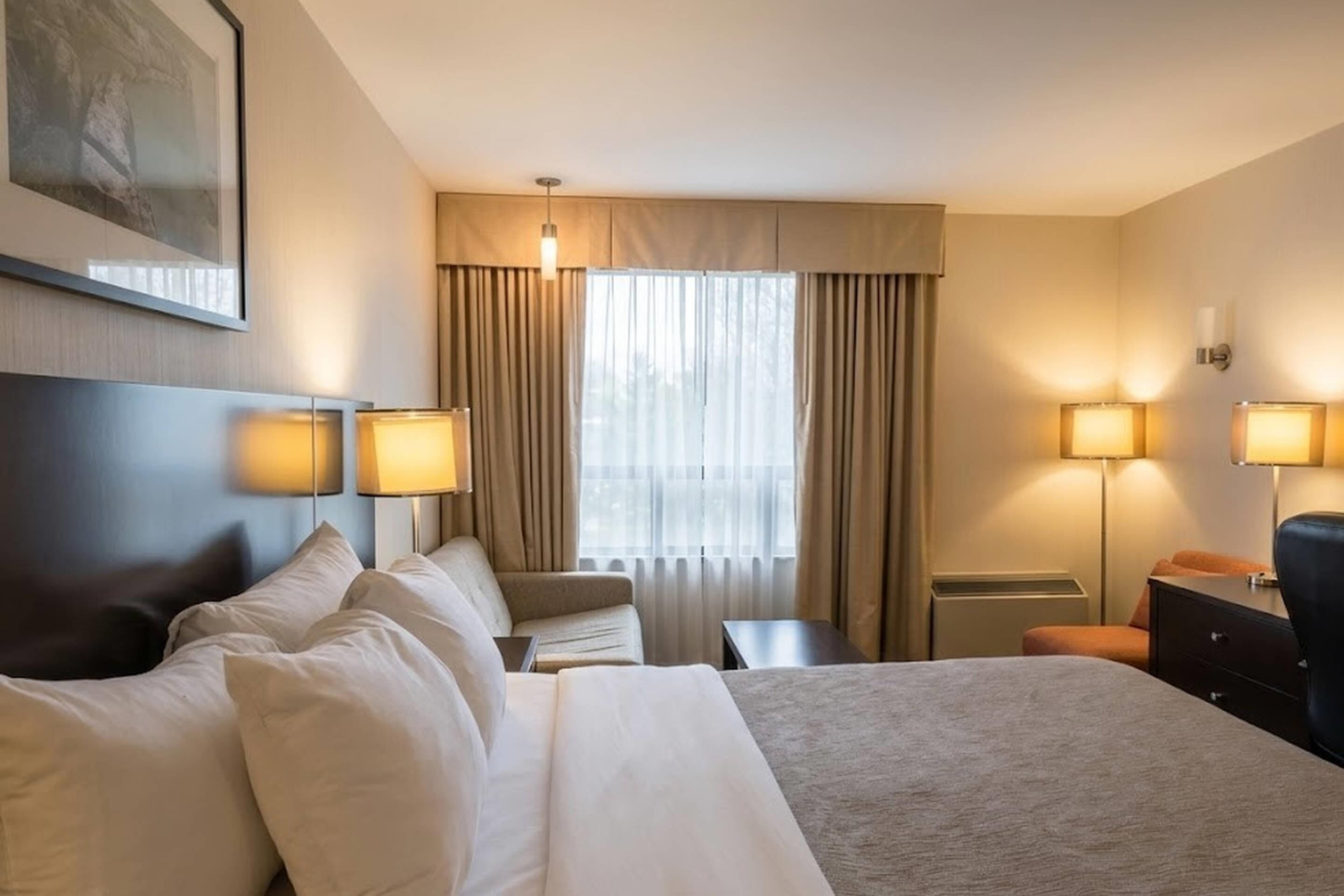 A hotel room with a large bed, two bedside tables, a chair, a desk, a lamp, and a window with curtains.