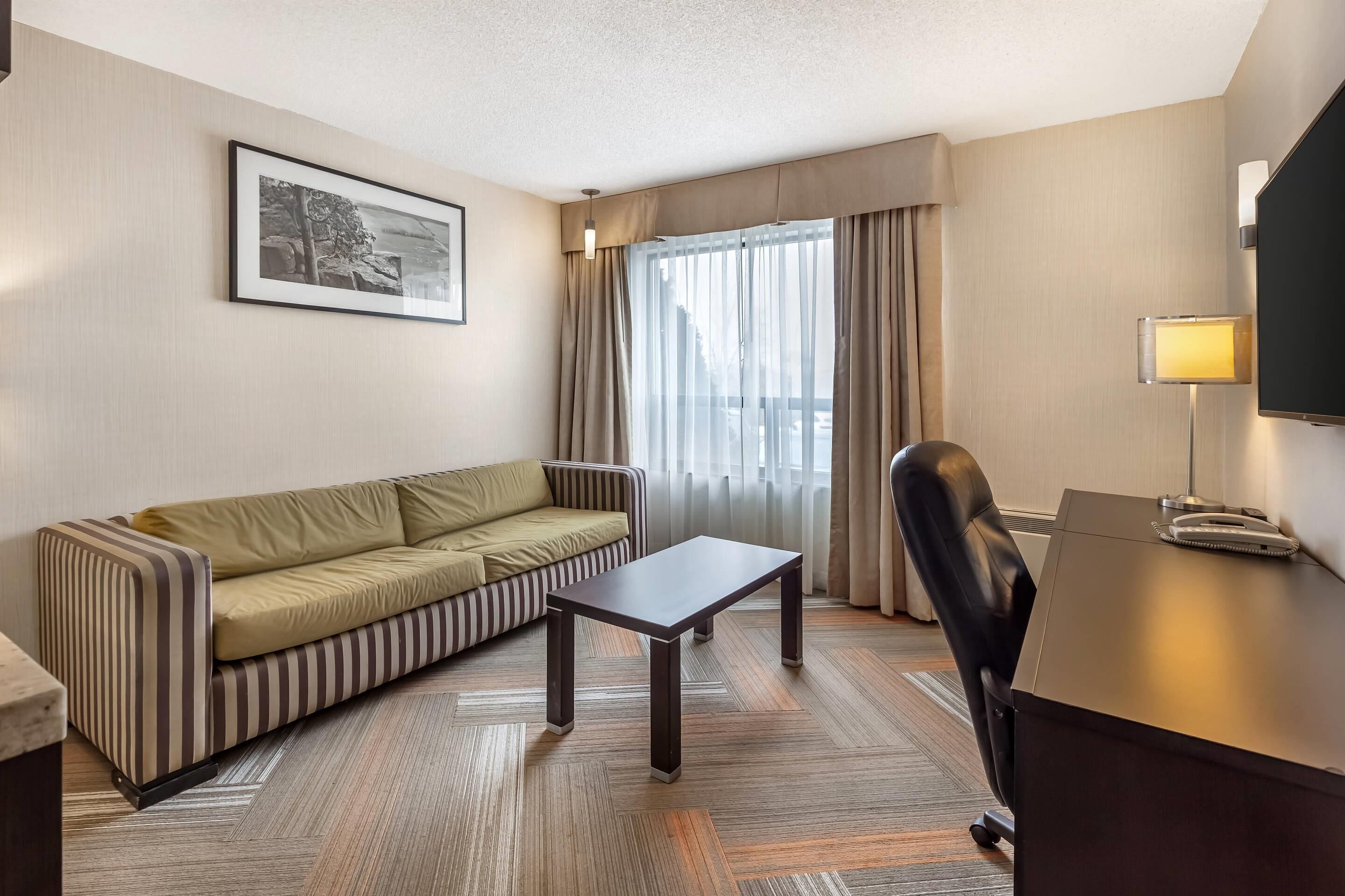 A hotel room with a striped sofa, a desk, a chair, a framed picture on the wall, and a window with curtains.