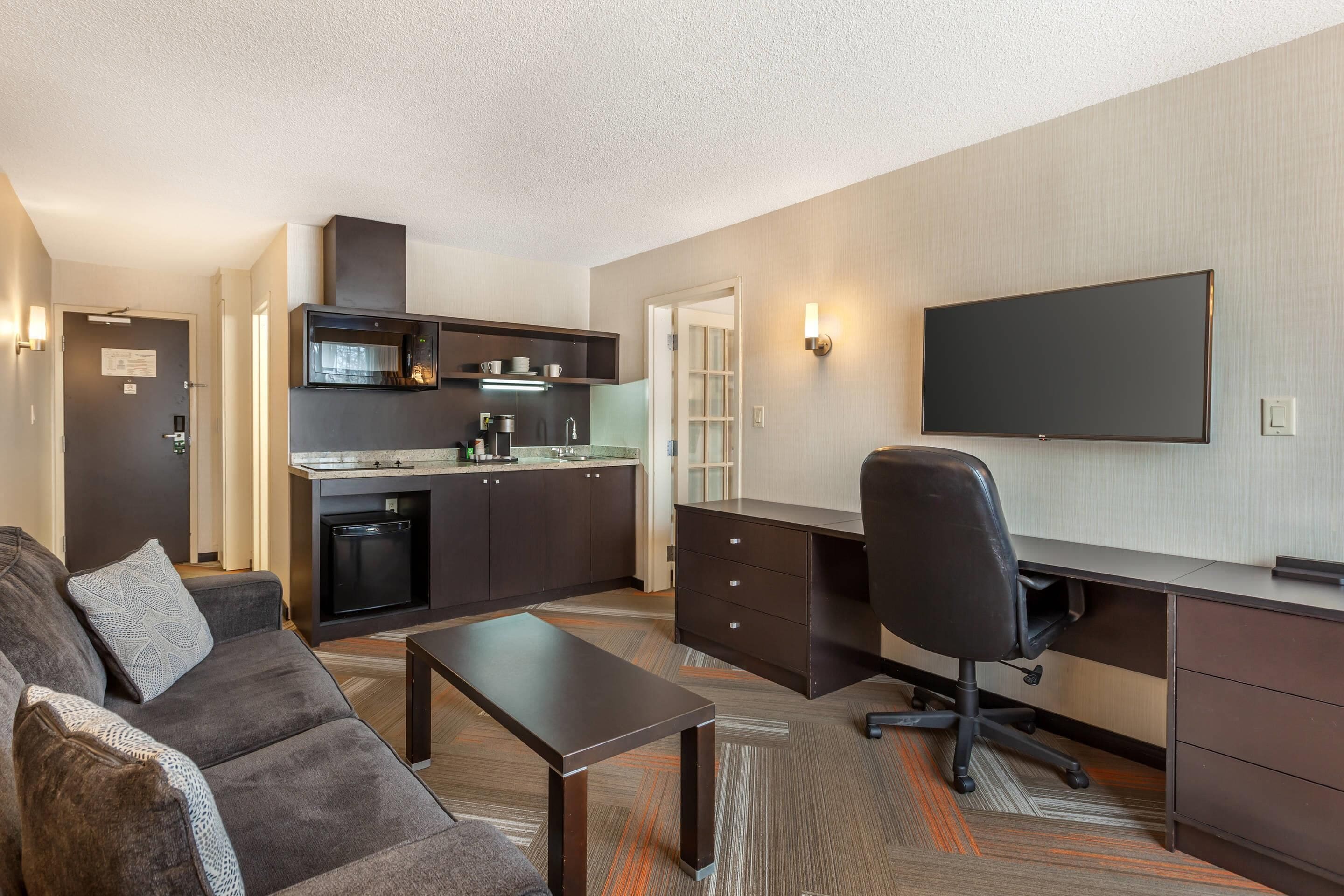 A compact living space with a kitchenette, a flat-screen TV, a sofa, a coffee table, and a desk with a chair.