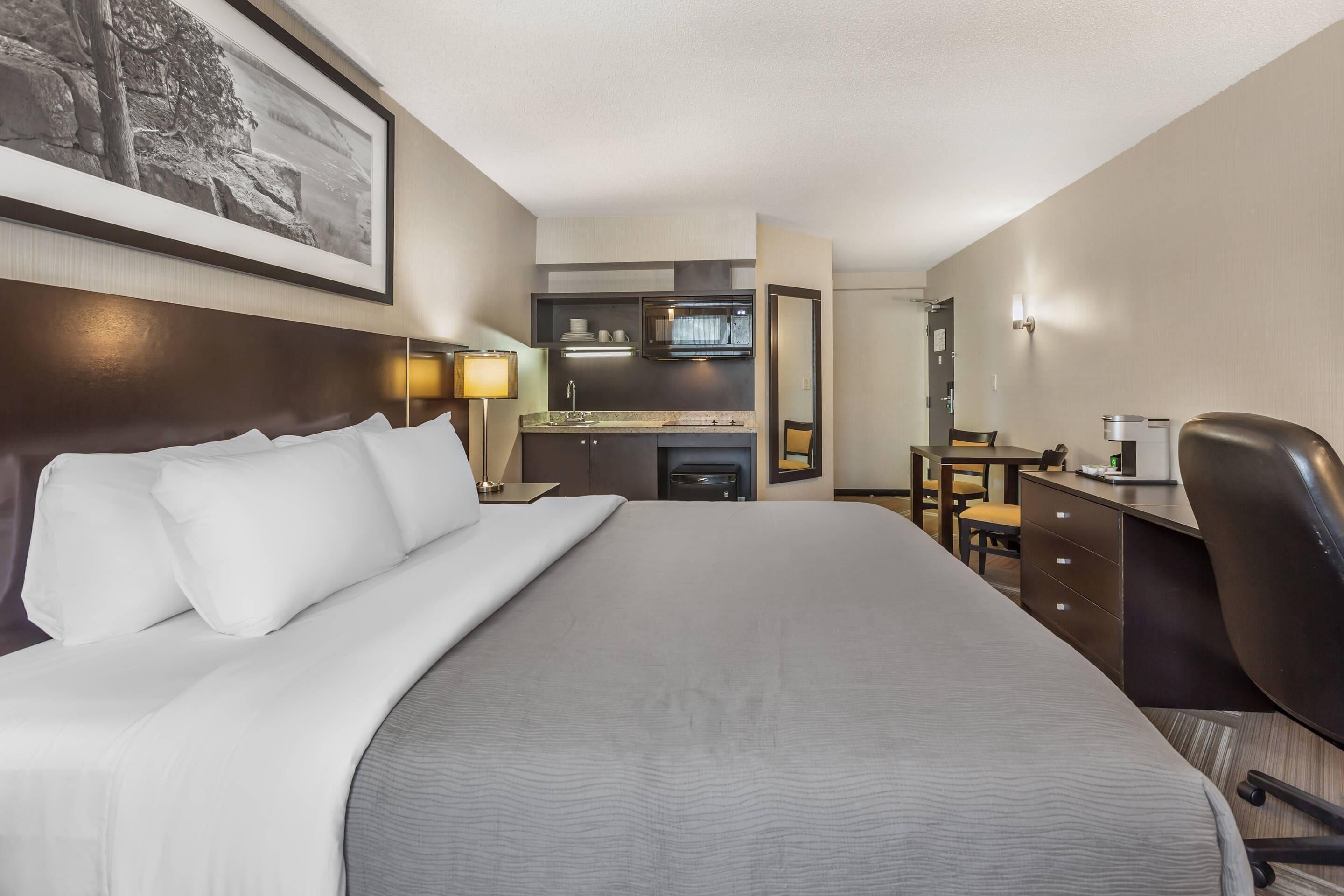 A hotel room with a large bed, a desk, a chair, a kitchenette, and a framed picture on the wall.