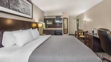A hotel room with a large bed, a desk, a chair, a kitchenette, and a framed picture on the wall.