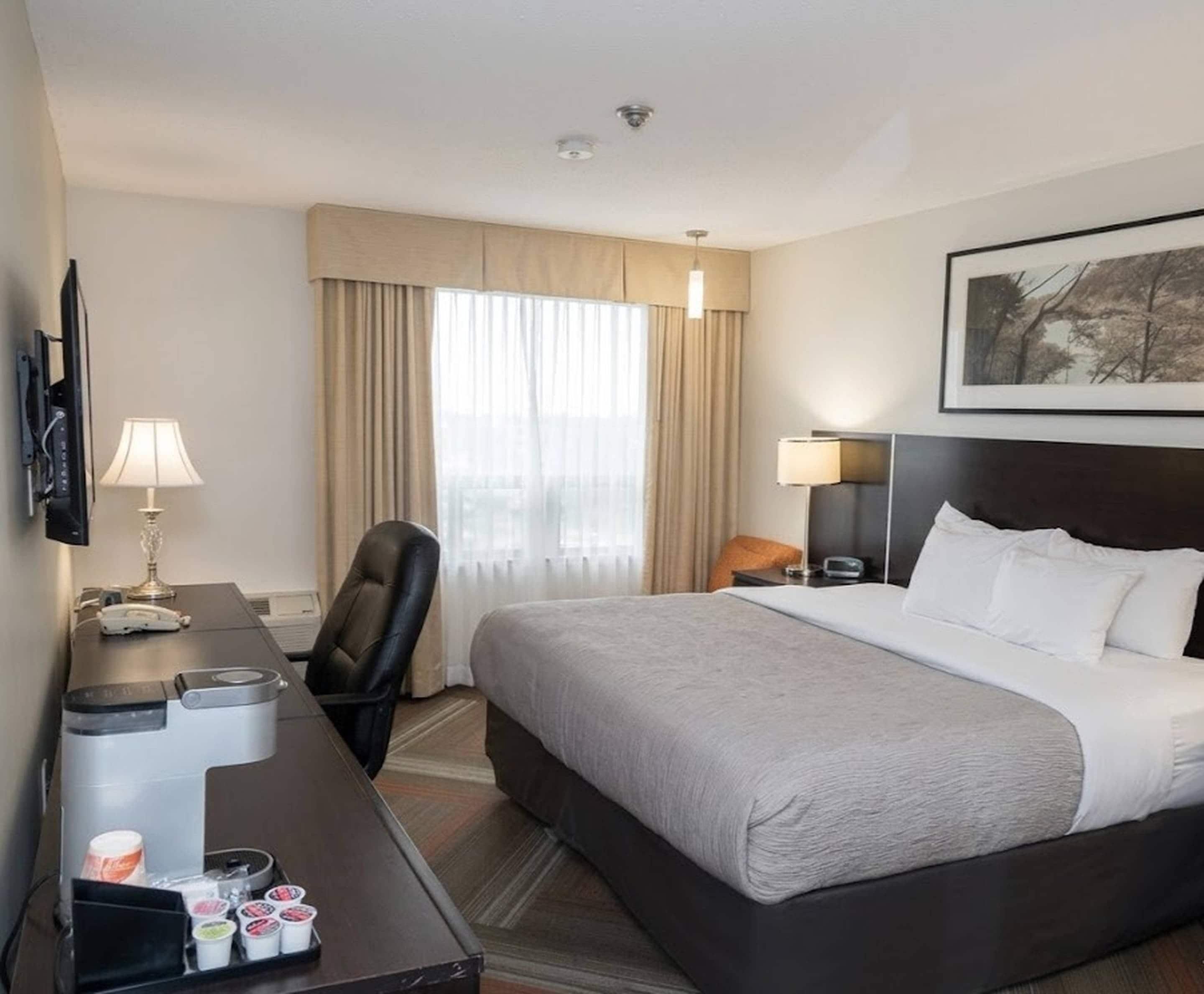 A hotel room with a bed, a desk with a phone and coffee maker, a chair, and a window with curtains.
