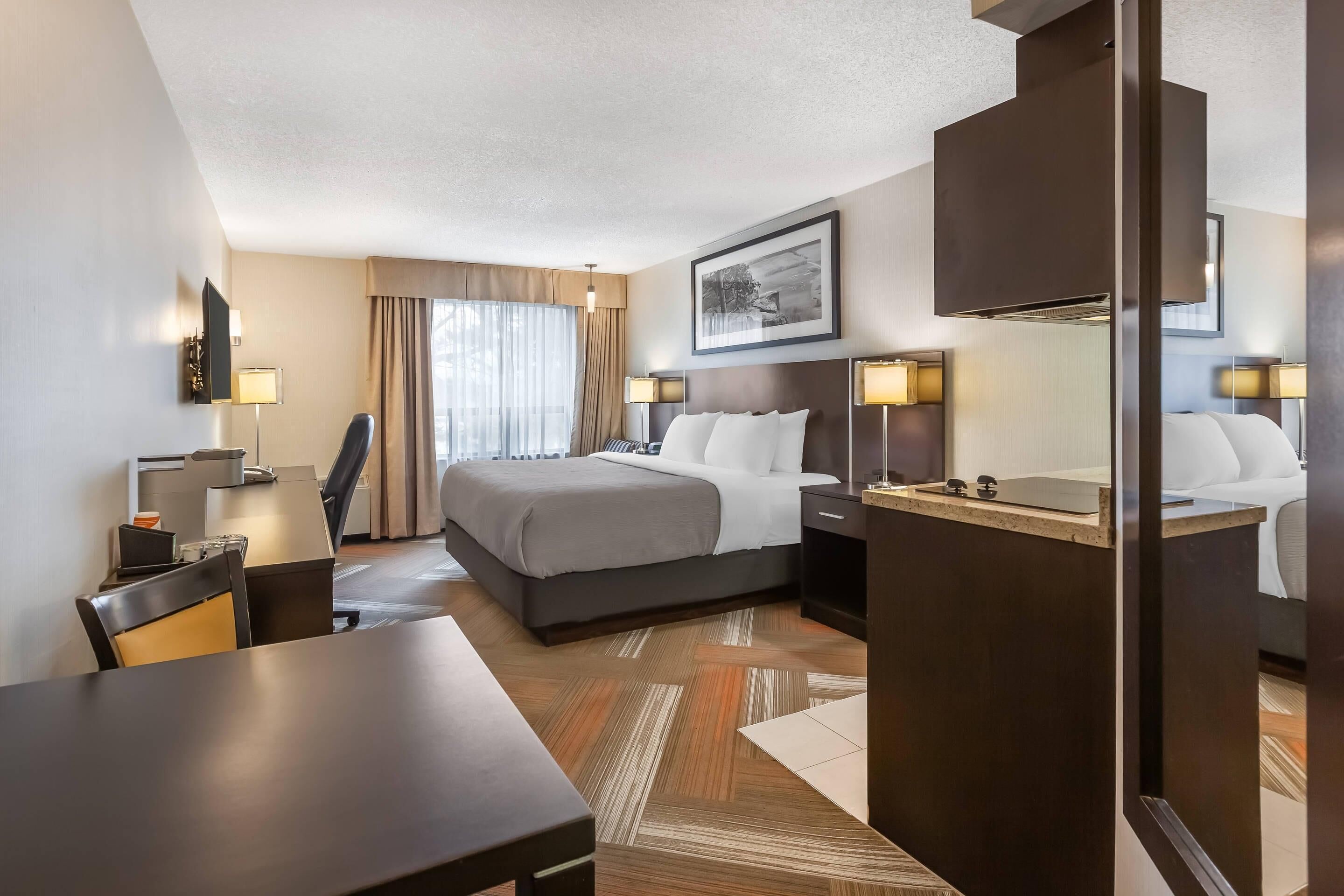 Habitación de hotel con cama, mesitas de noche, escritorio, silla, lámpara, espejo y ventana con cortinas.