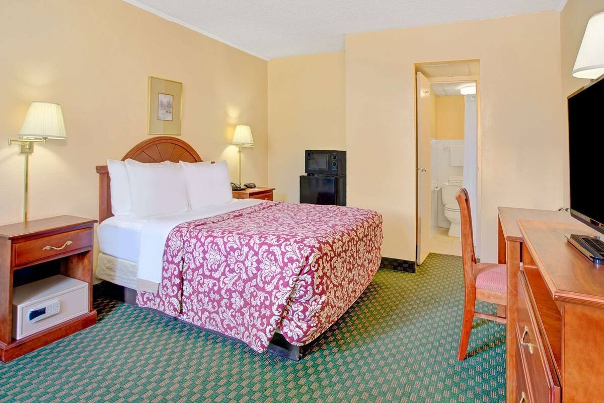 Habitación de hotel con cama, lámparas de noche, escritorio con silla, televisor y baño a la vista a través de una puerta abierta.