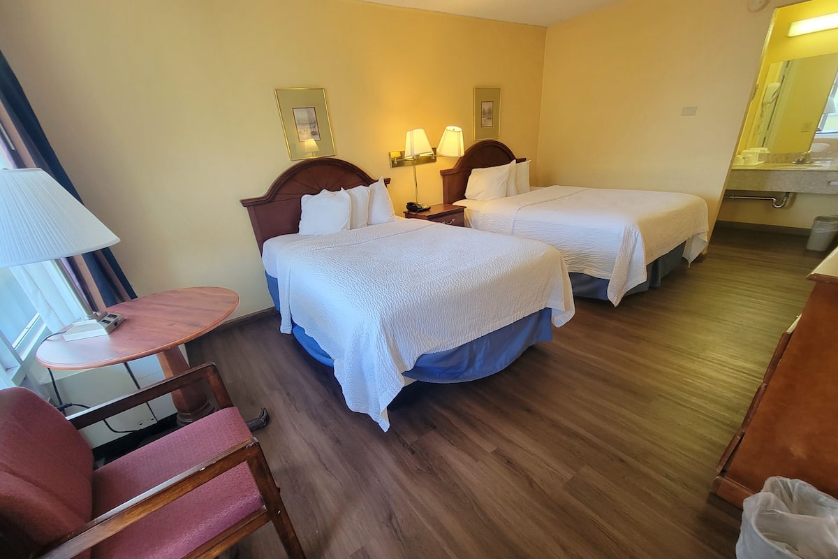 Habitación de hotel con dos camas, un escritorio, una silla y un baño visible al fondo.