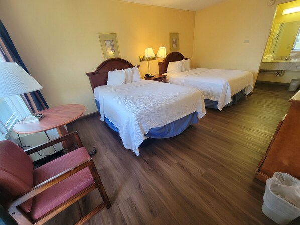 Quarto de hotel com duas camas, uma escrivaninha, uma cadeira e um banheiro ao fundo.