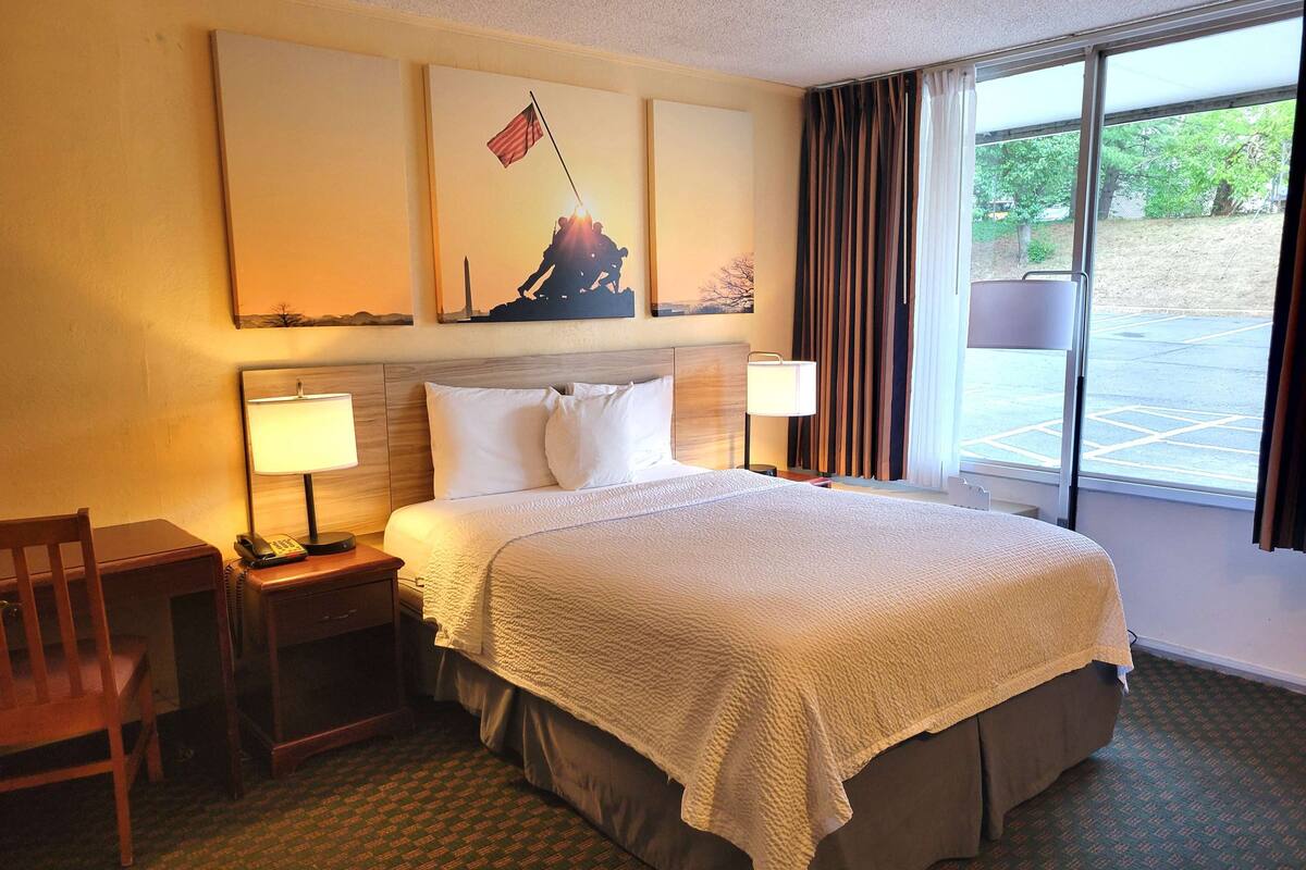 Habitación de hotel con una cama grande, dos mesitas de noche con lámparas, una silla de madera y un ventanal con cortinas.