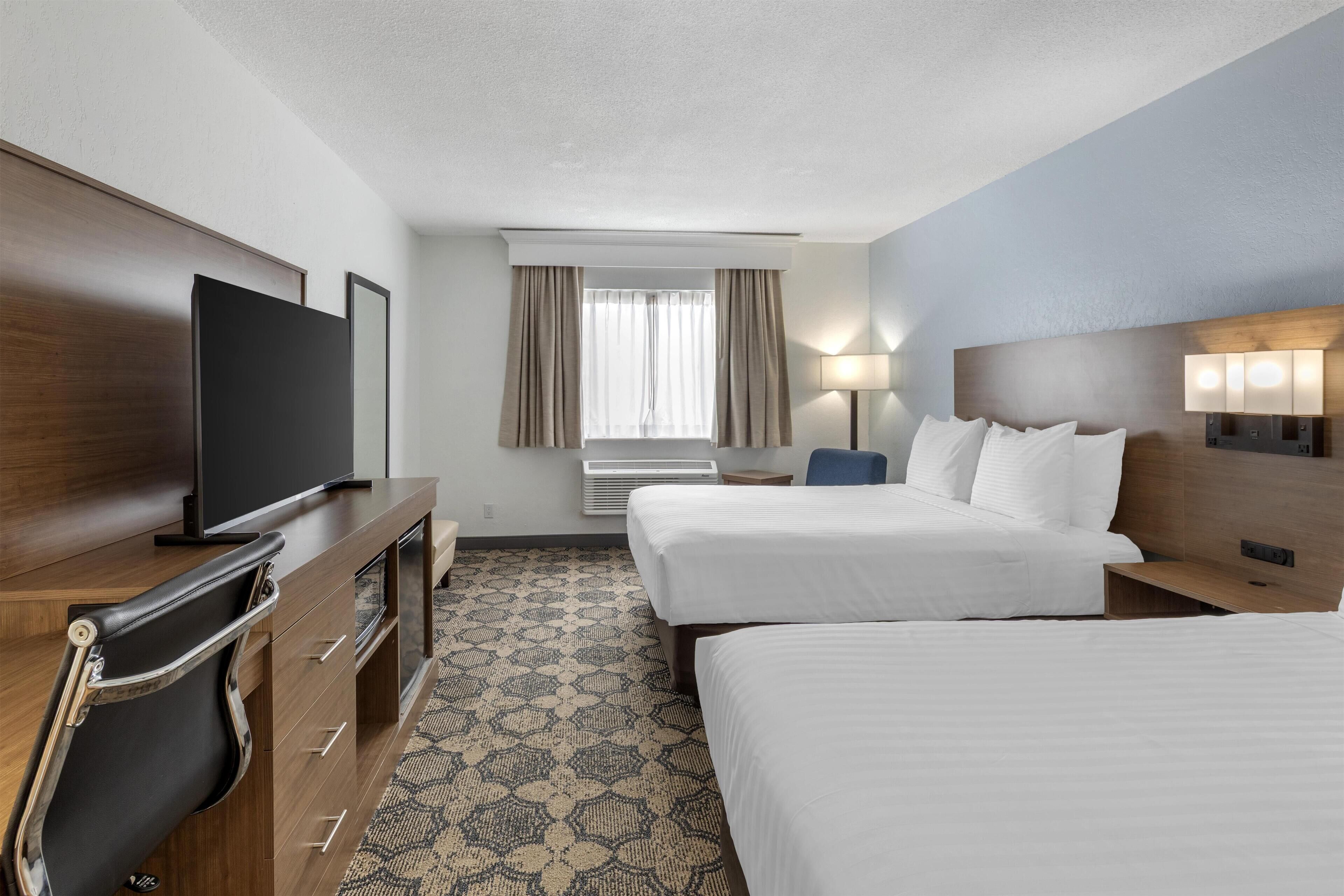 A hotel room with two beds, a TV, a desk with drawers, a chair, a window with curtains, and a patterned carpet.