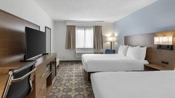 A hotel room with two beds, a TV, a desk with drawers, a chair, a window with curtains, and a patterned carpet.