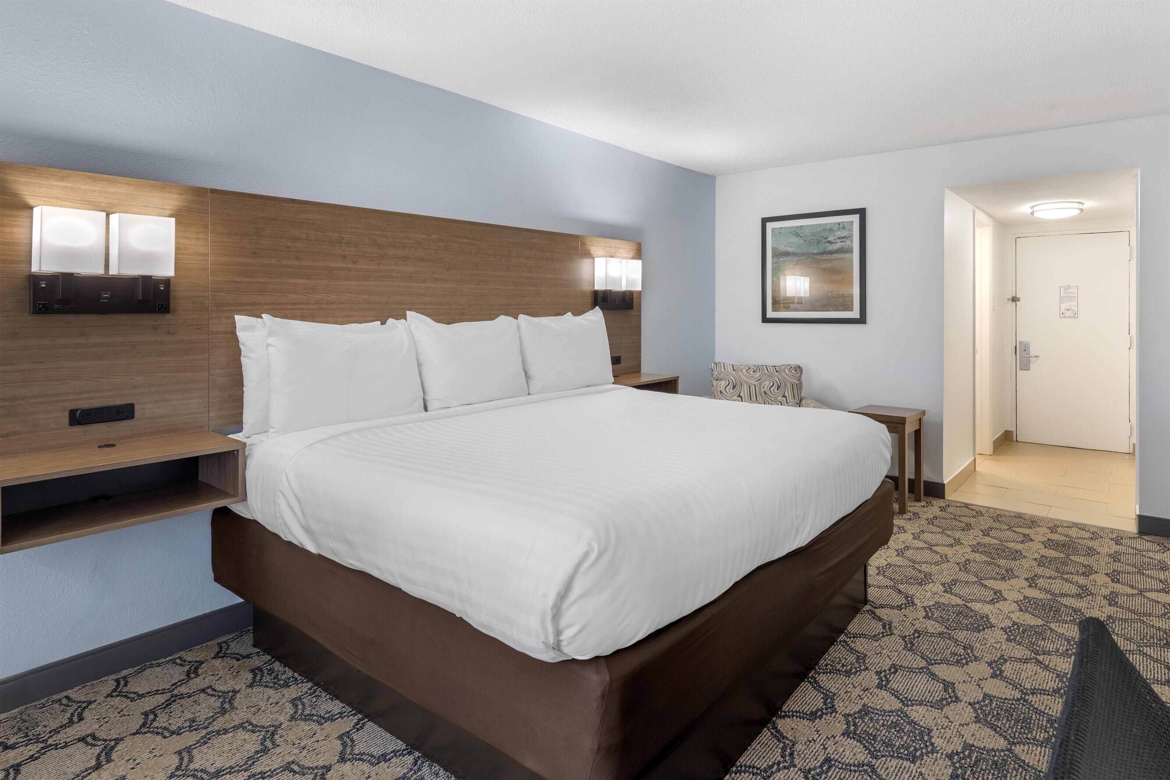 A hotel room with a large bed, a wooden headboard, a nightstand, a wall-mounted lamp, a framed picture, and a door leading to another room.