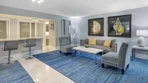 Un salon moderne avec un tapis bleu, deux fauteuils à motifs, une table basse blanche et un canapé gris agrémenté de coussins jaunes.
