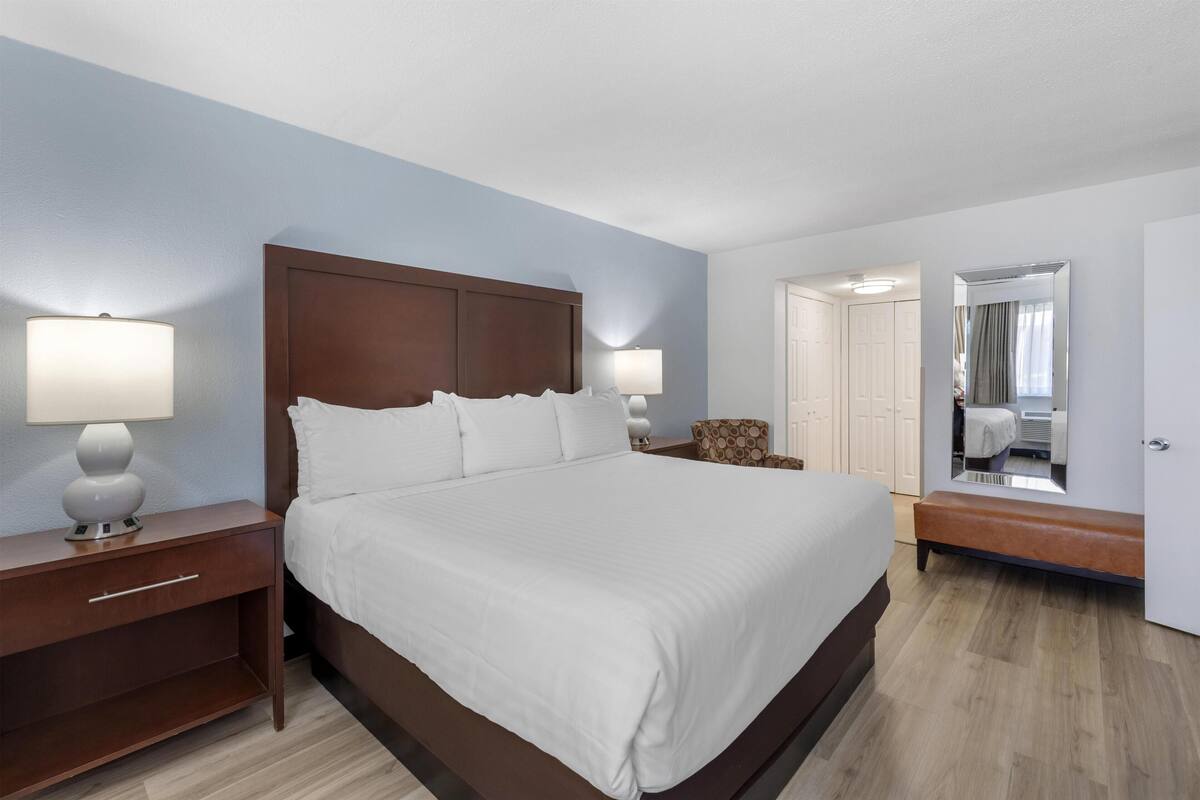A neatly made bed with white linens, a wooden headboard, two bedside tables with lamps, a bench, and a large mirror on the wall.