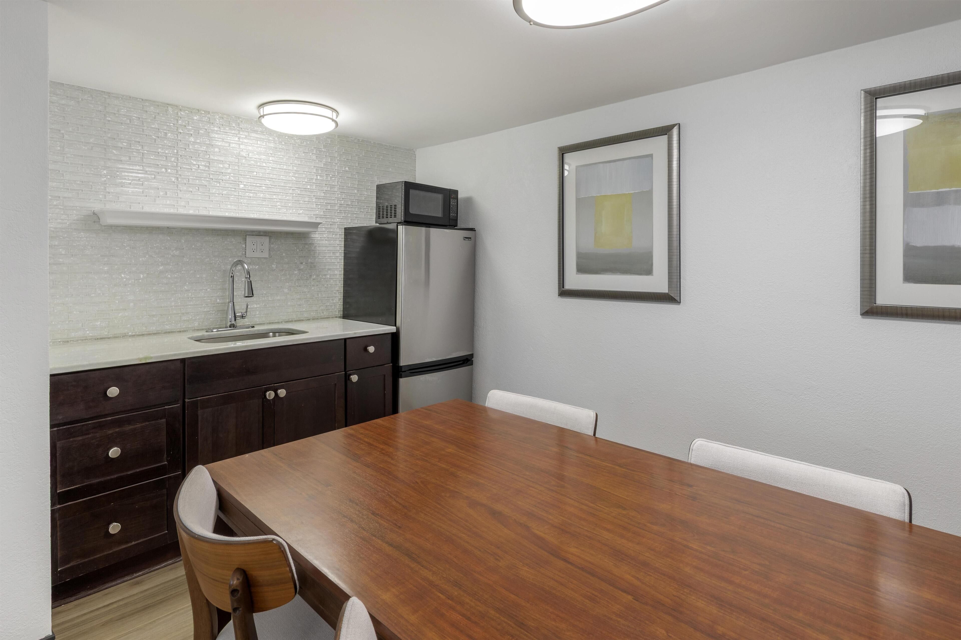 A modern kitchen with a wooden dining table, white walls, and a refrigerator.