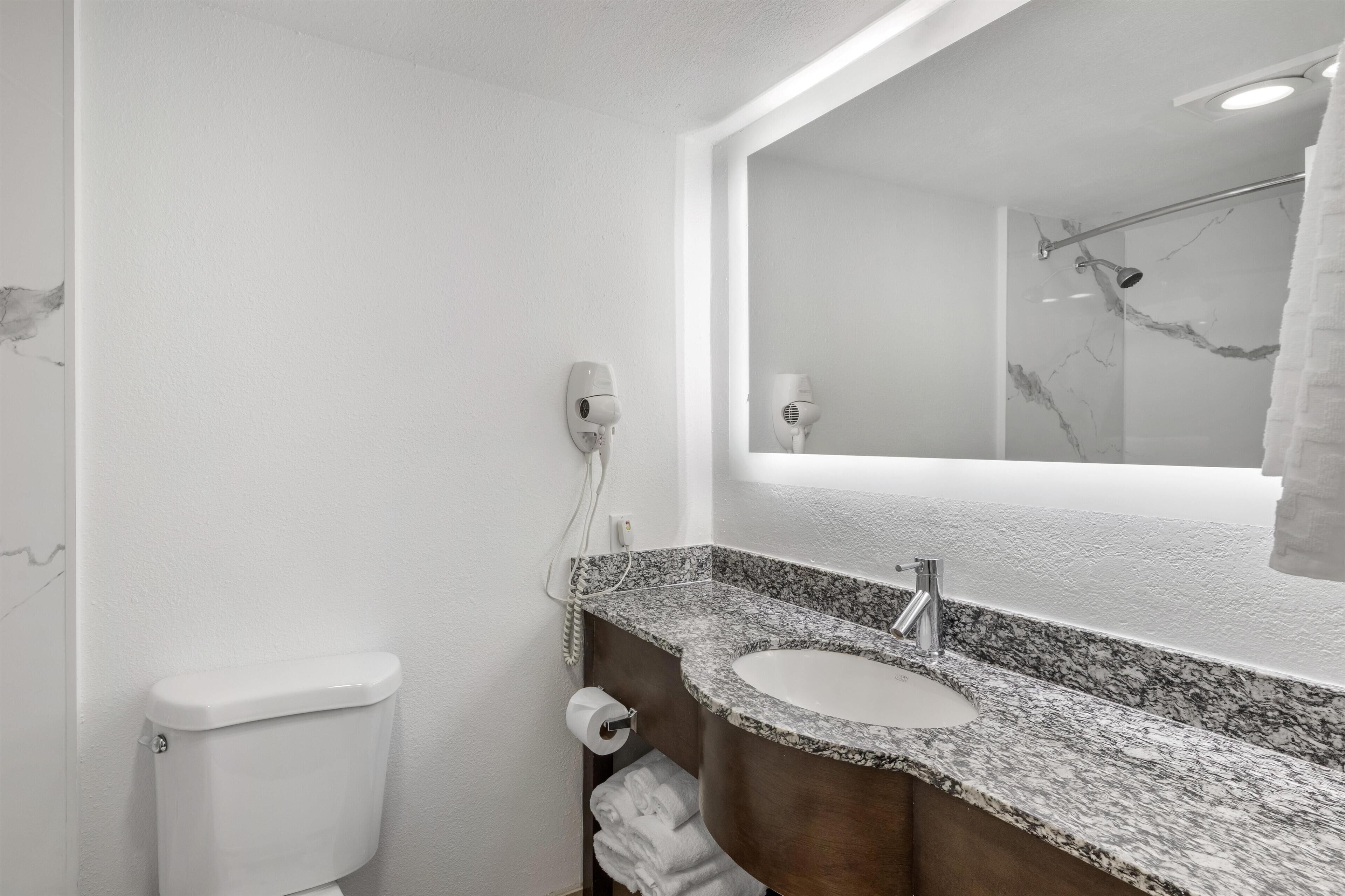 Un baño con inodoro blanco, lavabo con encimera de mármol y espejo sobre el lavabo.
