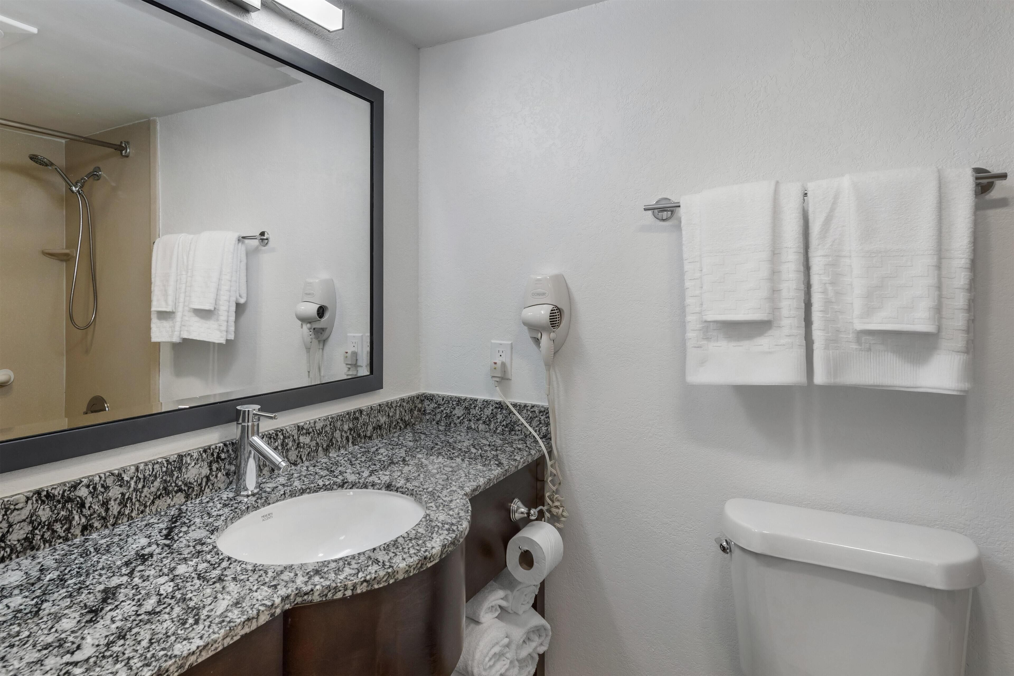 A bathroom with a granite countertop sink, a mirror, a towel rack with two towels, a hairdryer, and a toilet.