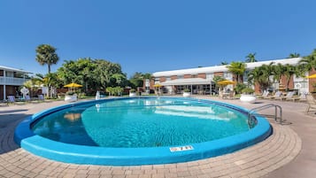 Une piscine dont la profondeur maximale est de 2,1 mètres, entourée de chaises longues et de parasols, avec un chemin pavé et des palmiers en arrière-plan.