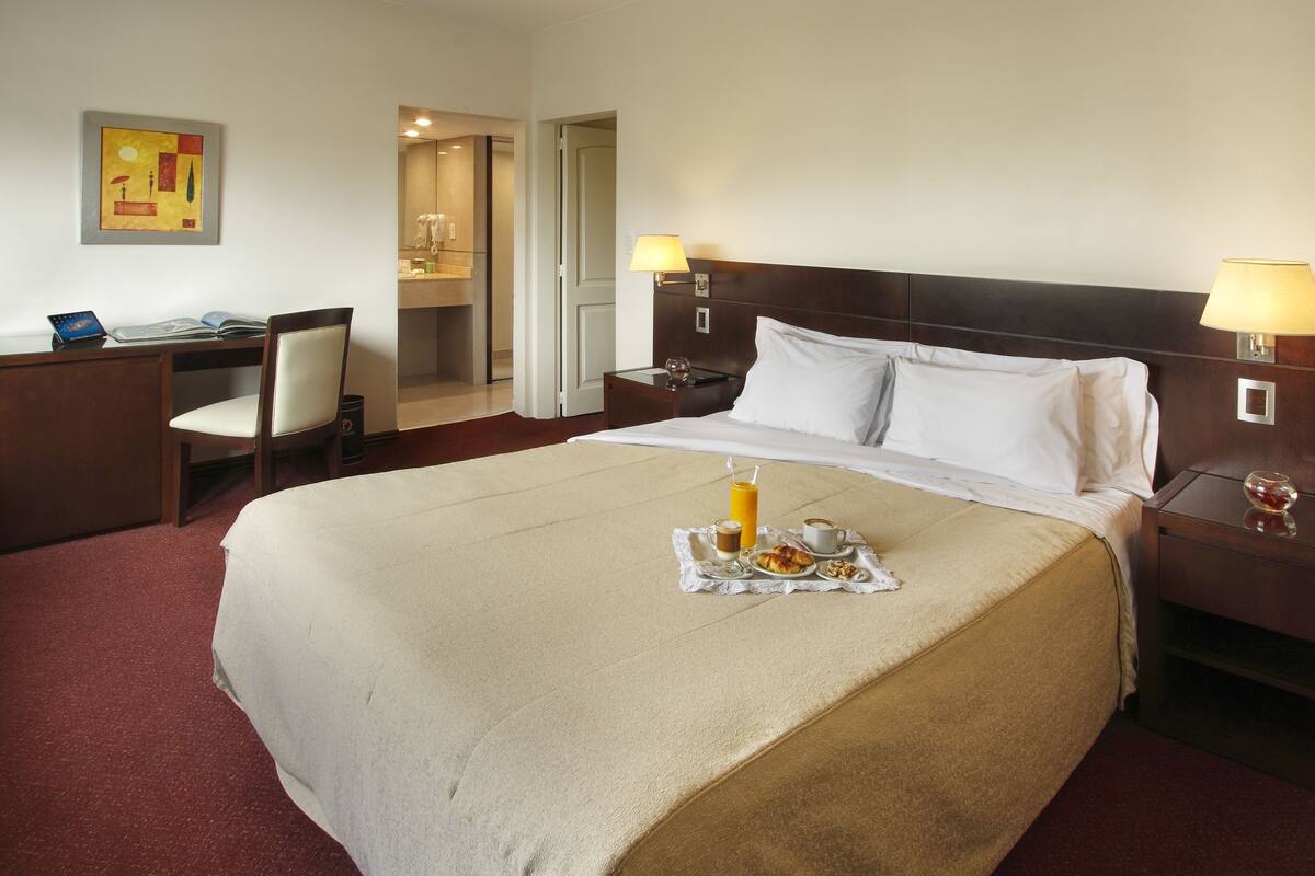 A hotel room with a large bed, two bedside tables with lamps, a desk with a chair, and a bathroom visible through an open door.