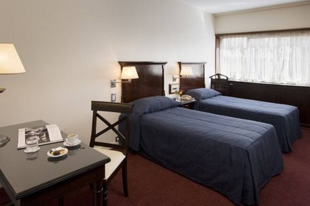 A hotel room with two single beds, a desk with a chair, a small table with a cup and a plate, and a window with curtains.