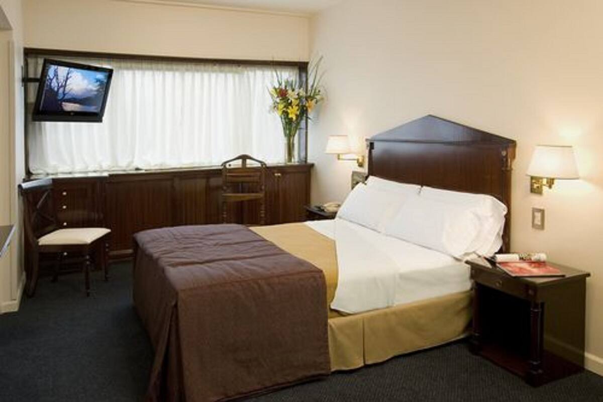 A hotel room with a bed, bedside tables, a desk, a chair, a TV, and a window with curtains.