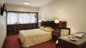 Habitación de hotel con cama, escritorio, silla, televisión, ventana con cortinas y lámpara.