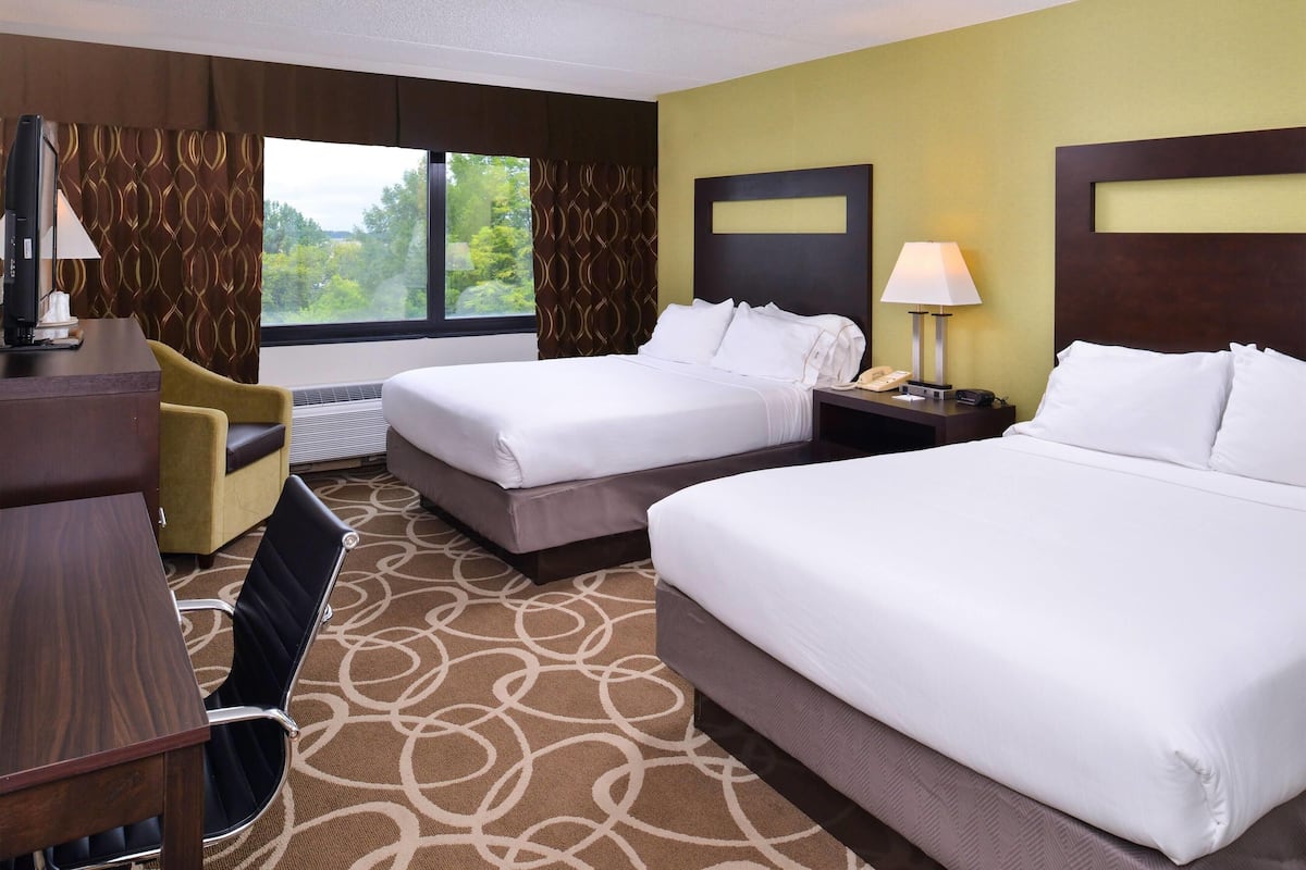 A hotel room with two beds, a desk, a chair, and a view of greenery outside.