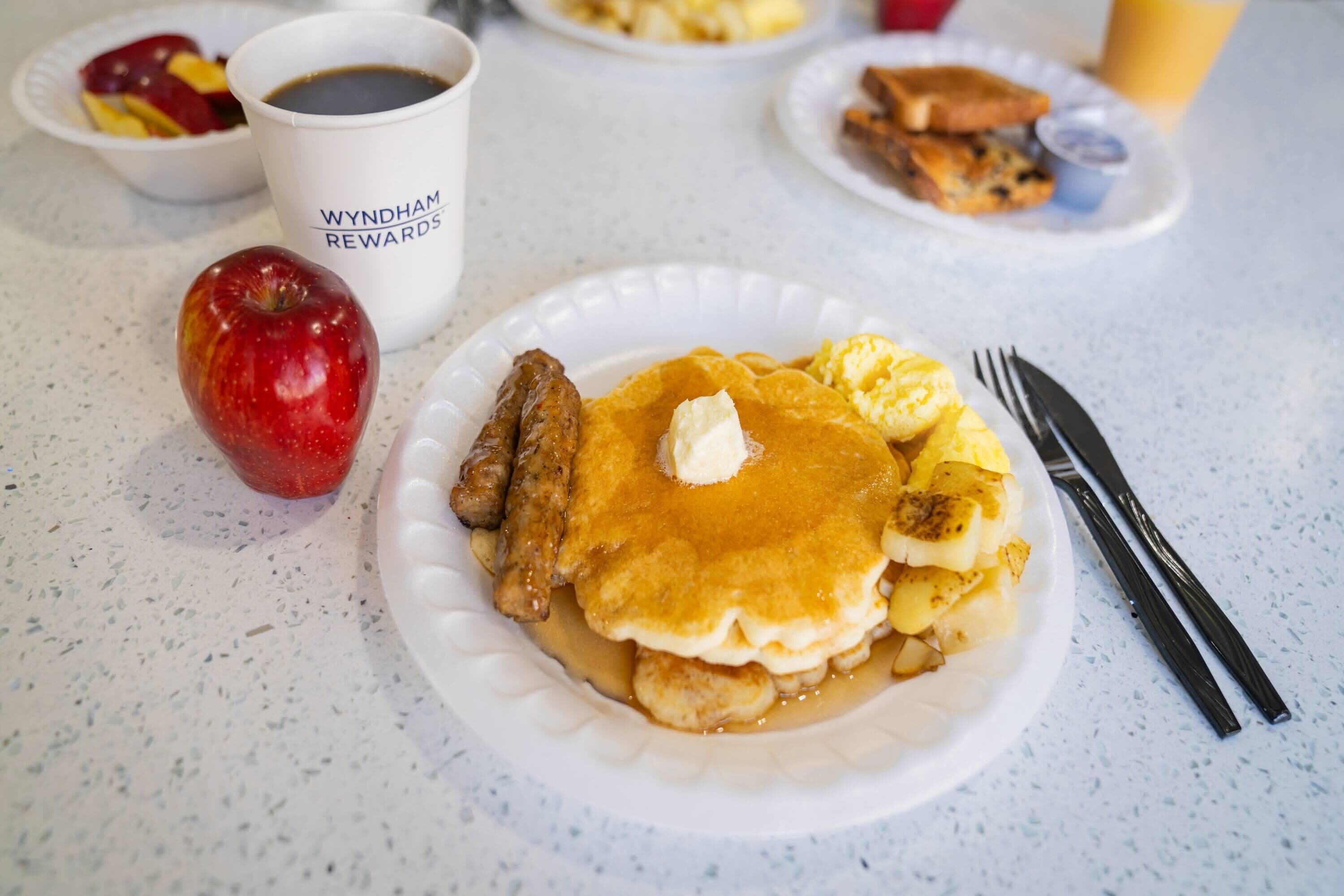 A breakfast plate with pancakes, sausage, and a red apple on a table.