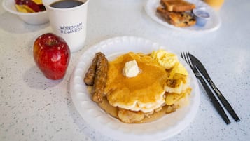 A breakfast plate with pancakes, sausage, and a red apple on a table.