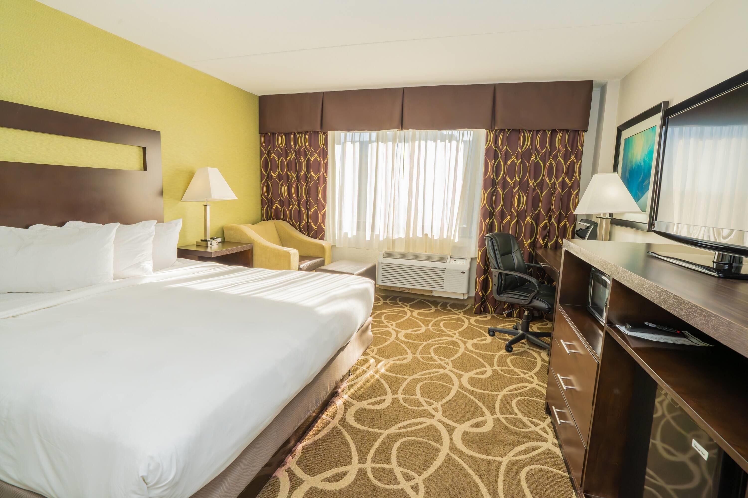 A hotel room with a large bed, a desk with a computer, a chair, a TV, and a window with curtains.