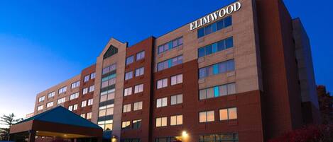 Ein mehrgeschossiges Hotel mit der Aufschrift 'ELIMWOOD' an der Fassade. Es verfügt über einen überdachten Eingang mit einer Garage, mehrere Fenster und einen Parkplatz mit Autos.