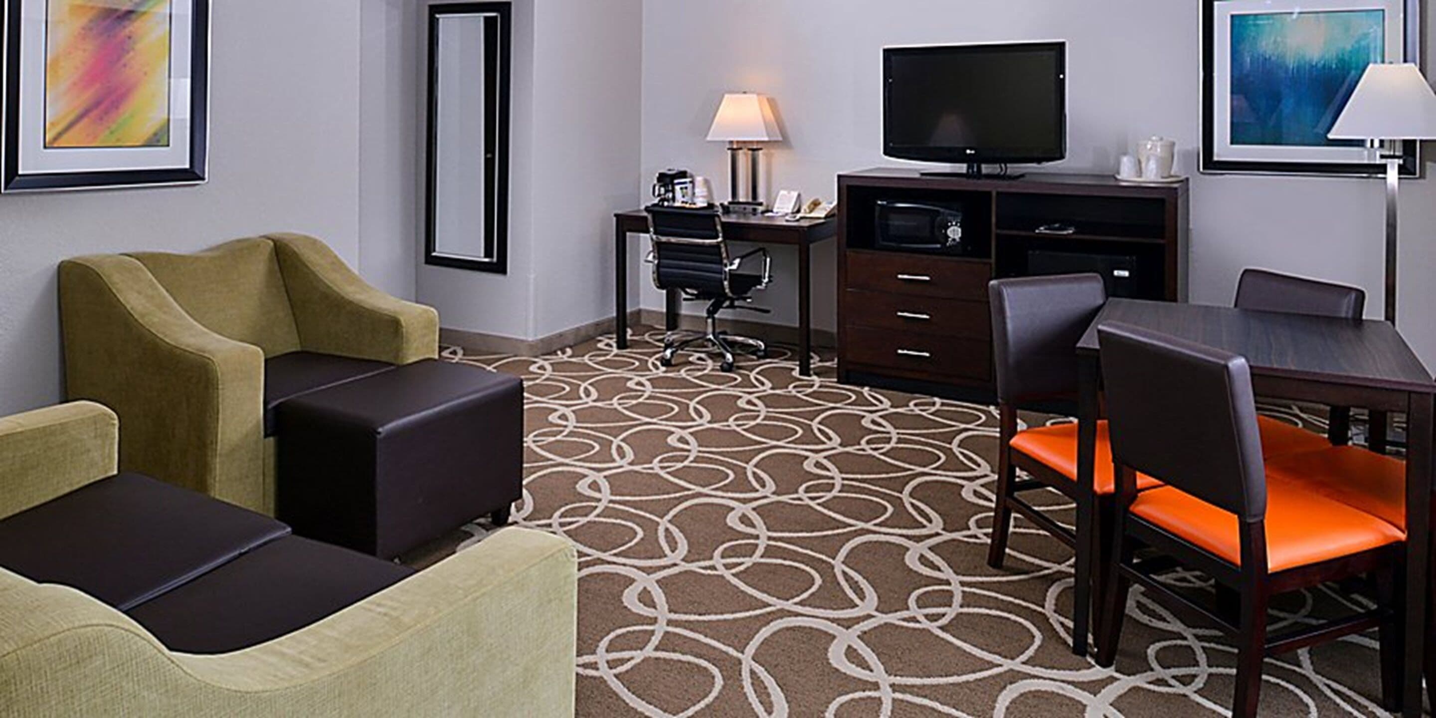 A hotel room with a sofa, a dining table with chairs, a TV, a desk with a lamp, and a patterned carpet.