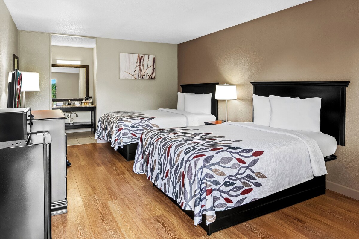 Une chambre d’hôtel avec deux lits, un bureau, un miroir et une salle de bain visible à travers une porte ouverte.