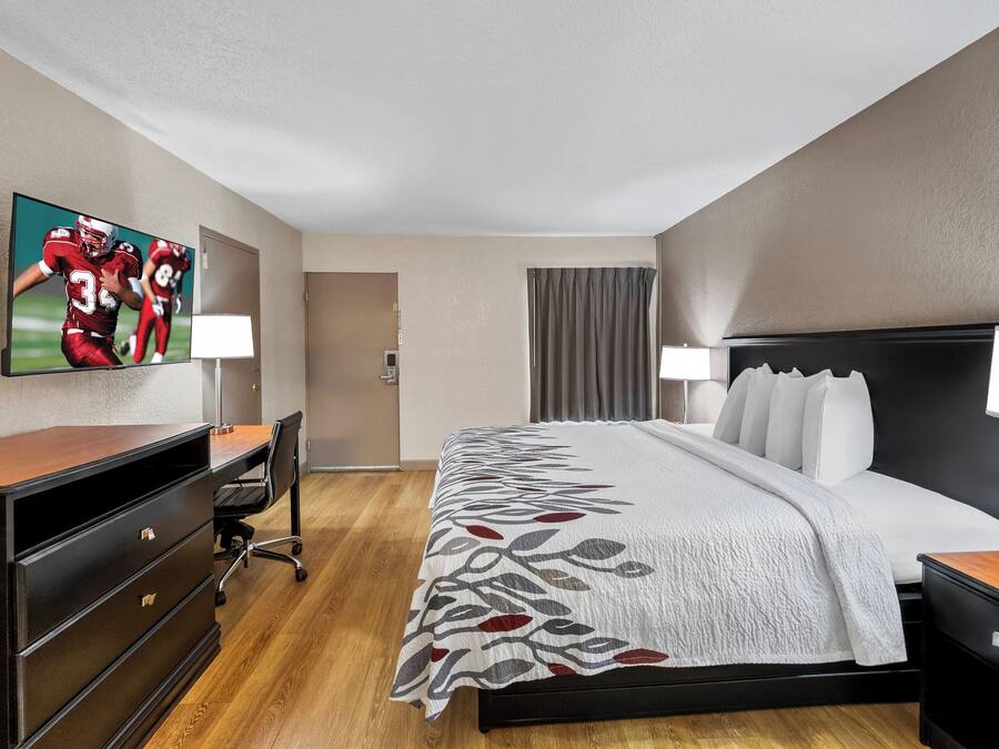 Ein Hotelzimmer mit einem großen Bett, einem Schreibtisch mit Stuhl, einem Wandfernseher und einem Fenster mit Vorhängen.