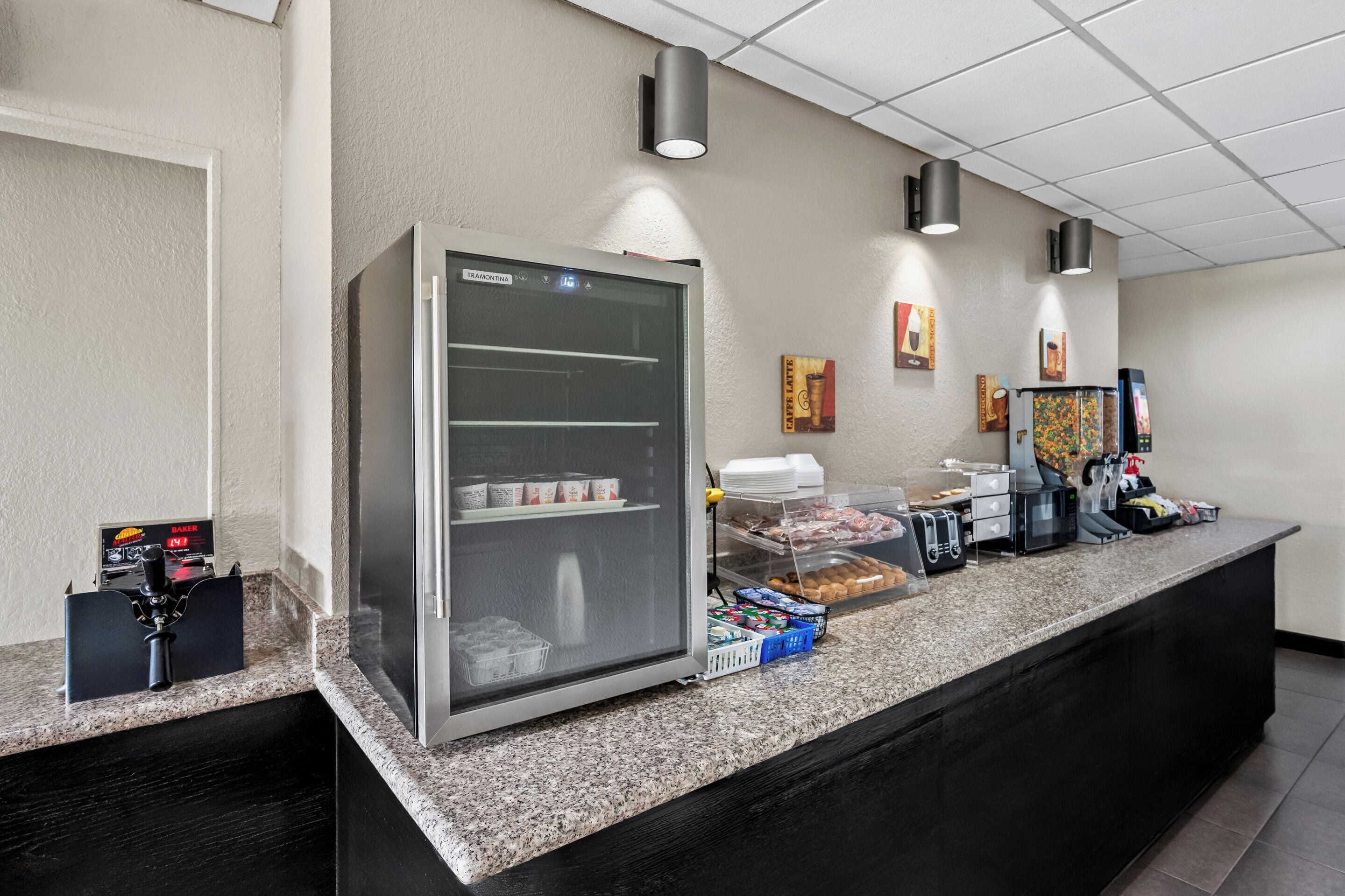 Un área de desayuno autoservicio con un refrigerador, máquinas de café y una variedad de alimentos.