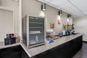 A self-service breakfast area with a refrigerated display case, coffee machines, and a variety of food items.