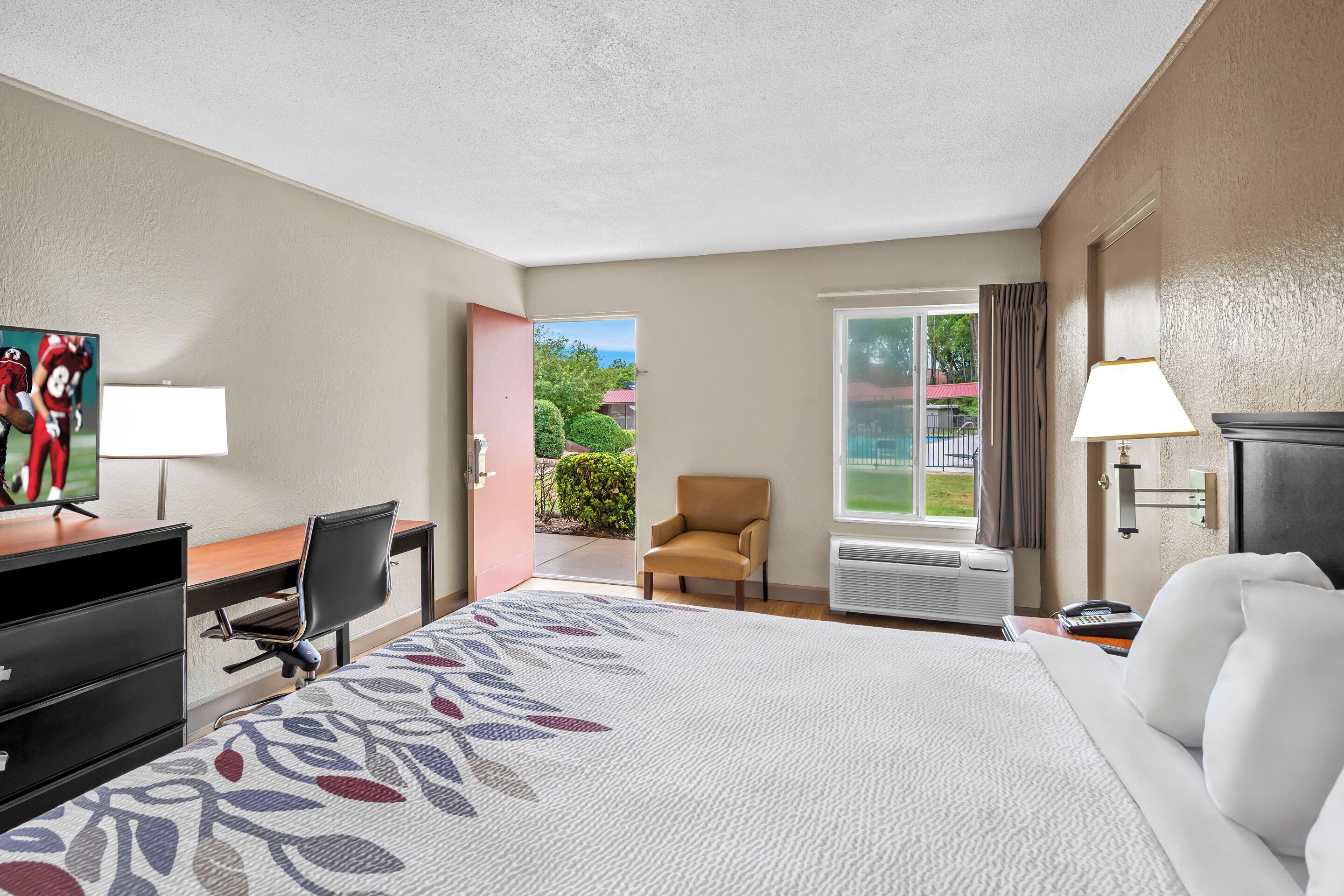 A hotel room with a bed, desk, chair, and a view of the outdoors.