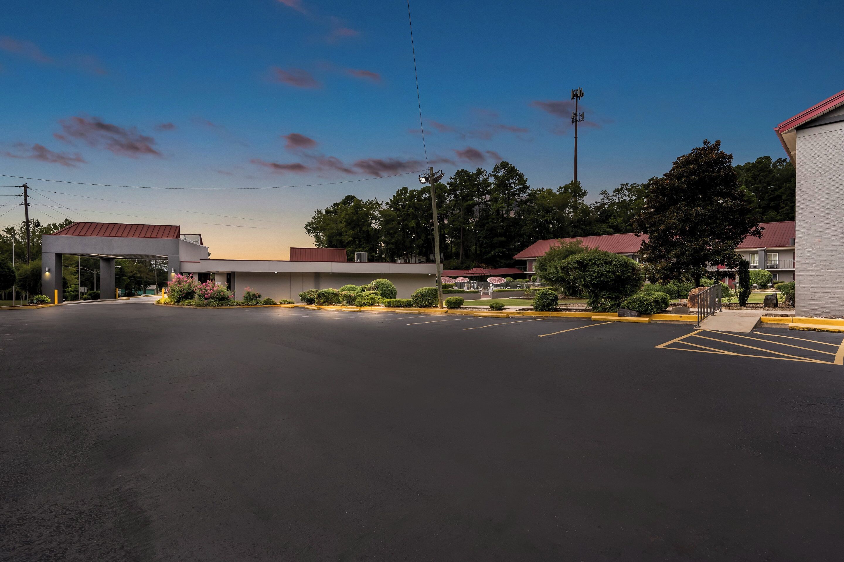 A parking lot with a building that has a red roof and a signboard. There are trees and bushes around the building.
