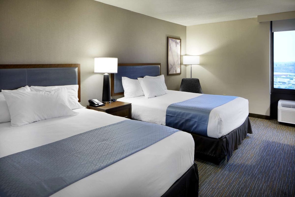 A hotel room with two beds, a desk with a lamp, a chair, and a large window with a view.