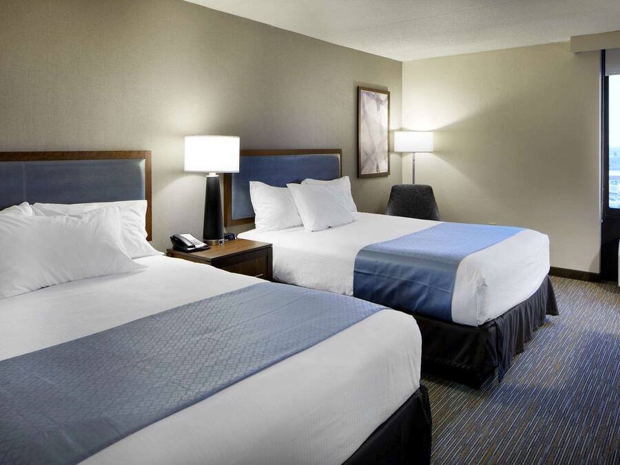 A hotel room with two beds, a desk with a lamp, a chair, and a large window with a view.