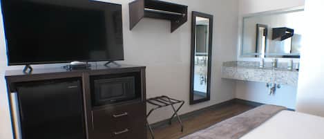 A hotel room with a flat-screen TV, a microwave, a small table, a mirror, and a bathroom with a sink and mirror.