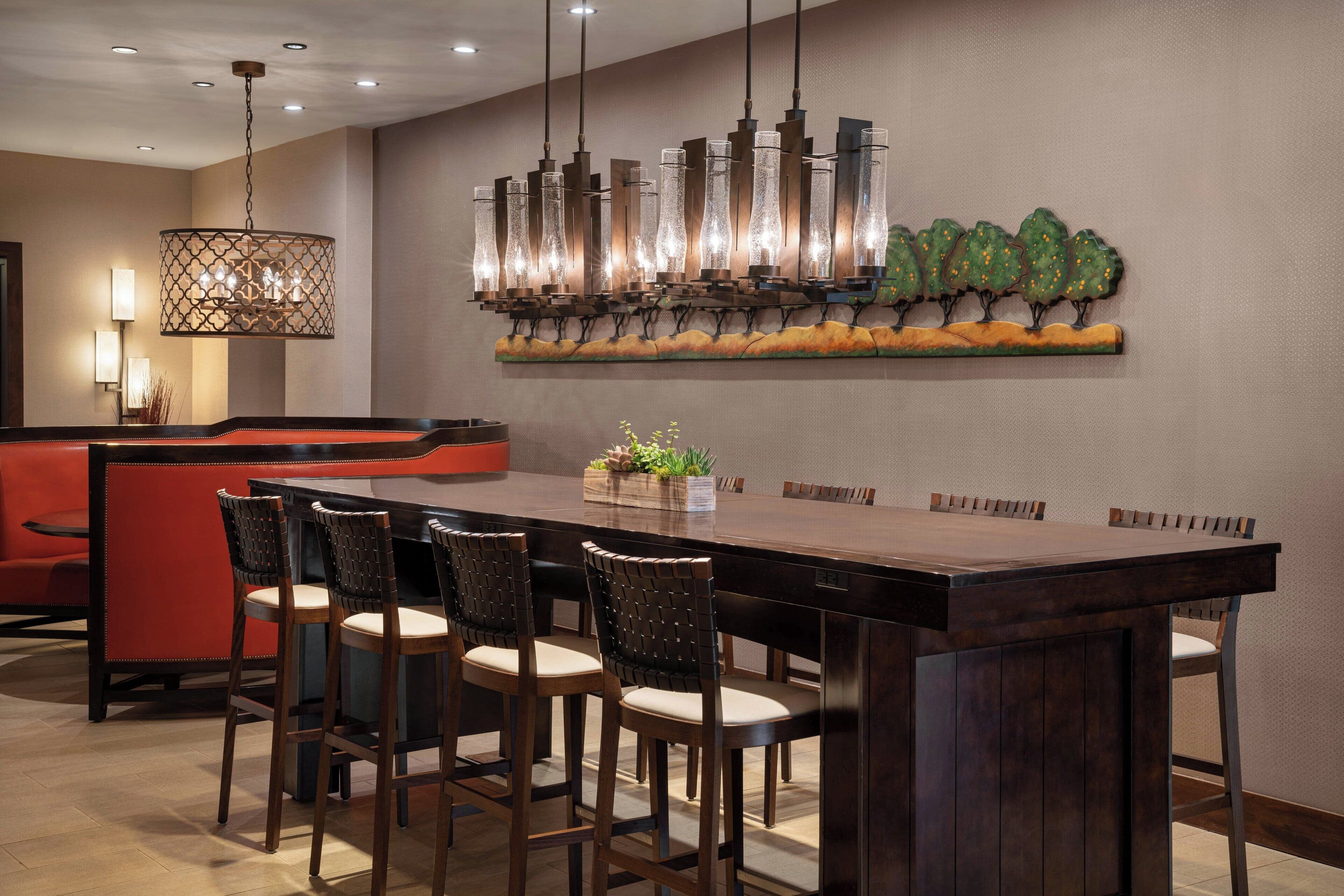 Un moderno vestíbulo de hotel con un largo mostrador de recepción de madera, varios taburetes de bar, una zona de estar roja y una pared decorativa con motivos de árboles.