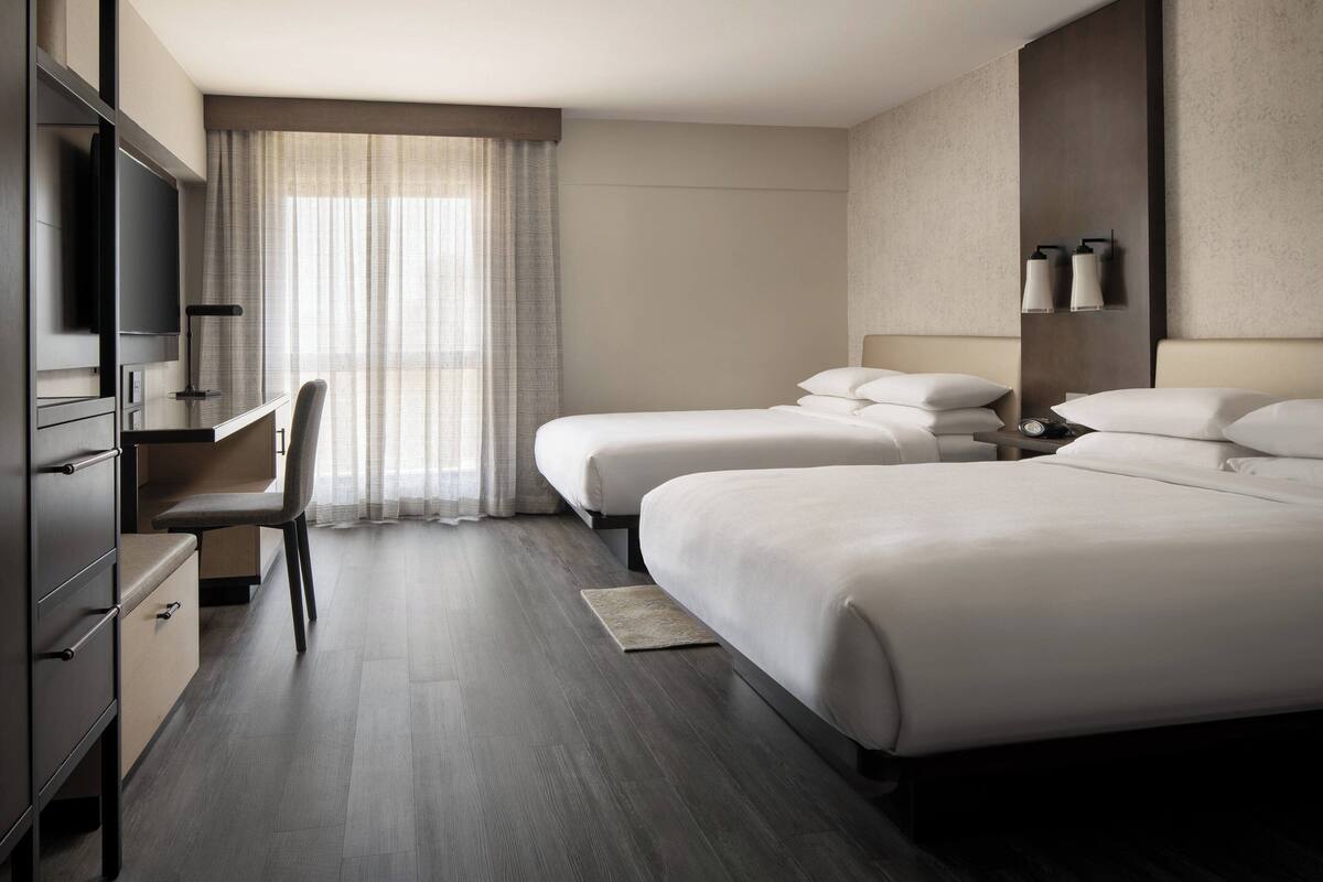 A hotel room with two beds, a desk, a TV, and a large window with curtains.