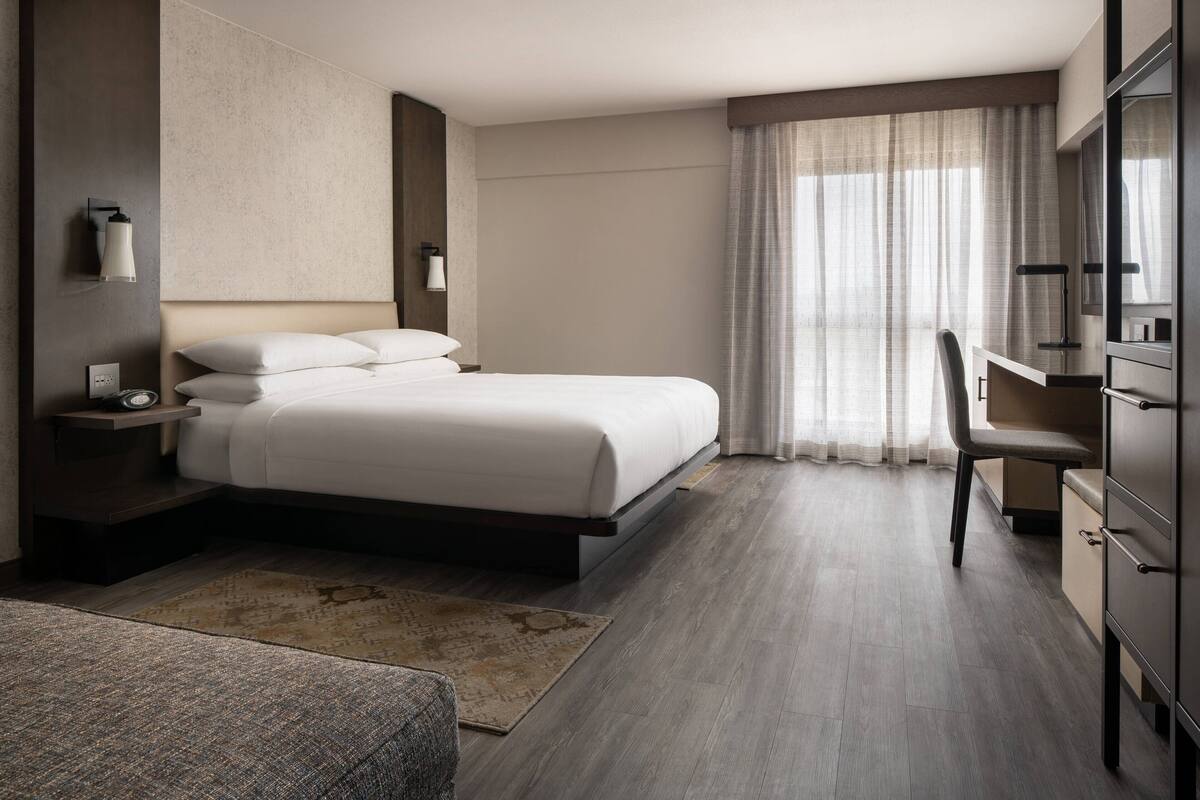 A modern hotel room with a large bed, a desk, and a chair. There is a small rug on the floor and a wall-mounted lamp.